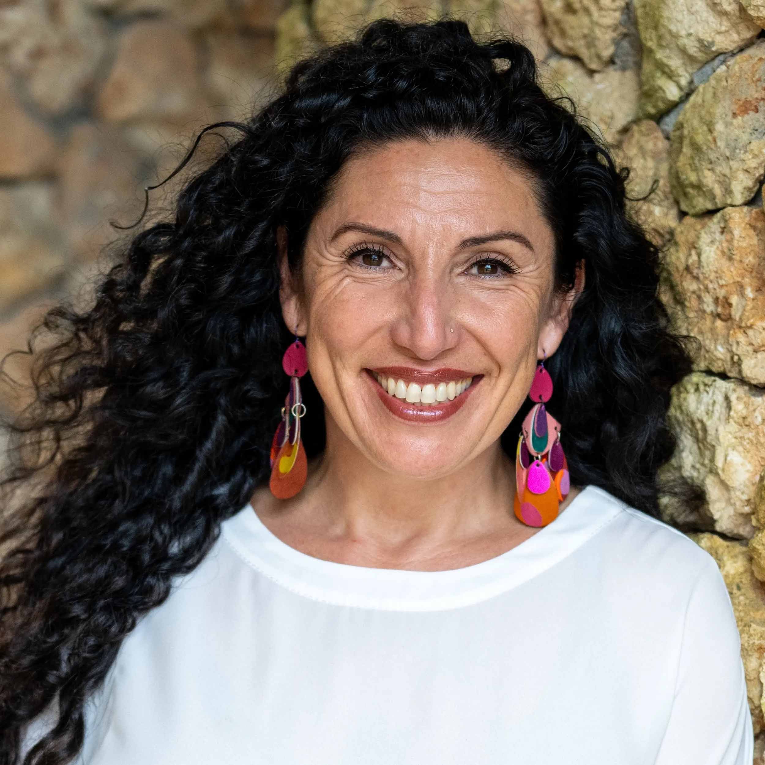 Maritza Schafer smiles at the camera while standing near a stone wall, wearing a white top and colorful dangling earrings.