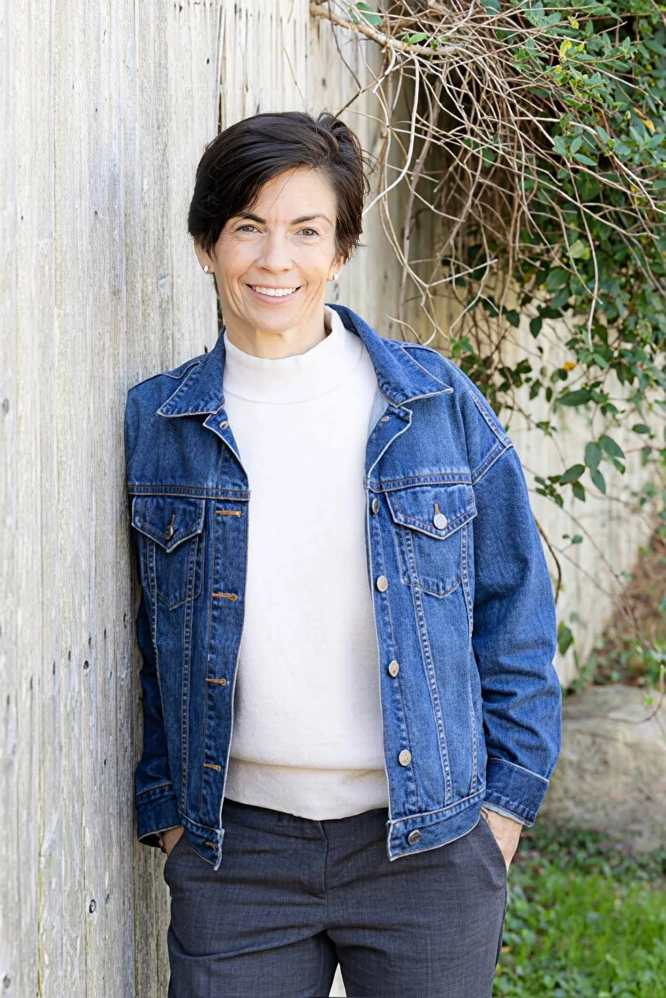 Woman with short dark hair wearing a denim jacket over a white sweater, leaning against a weathered wooden wall and smiling.