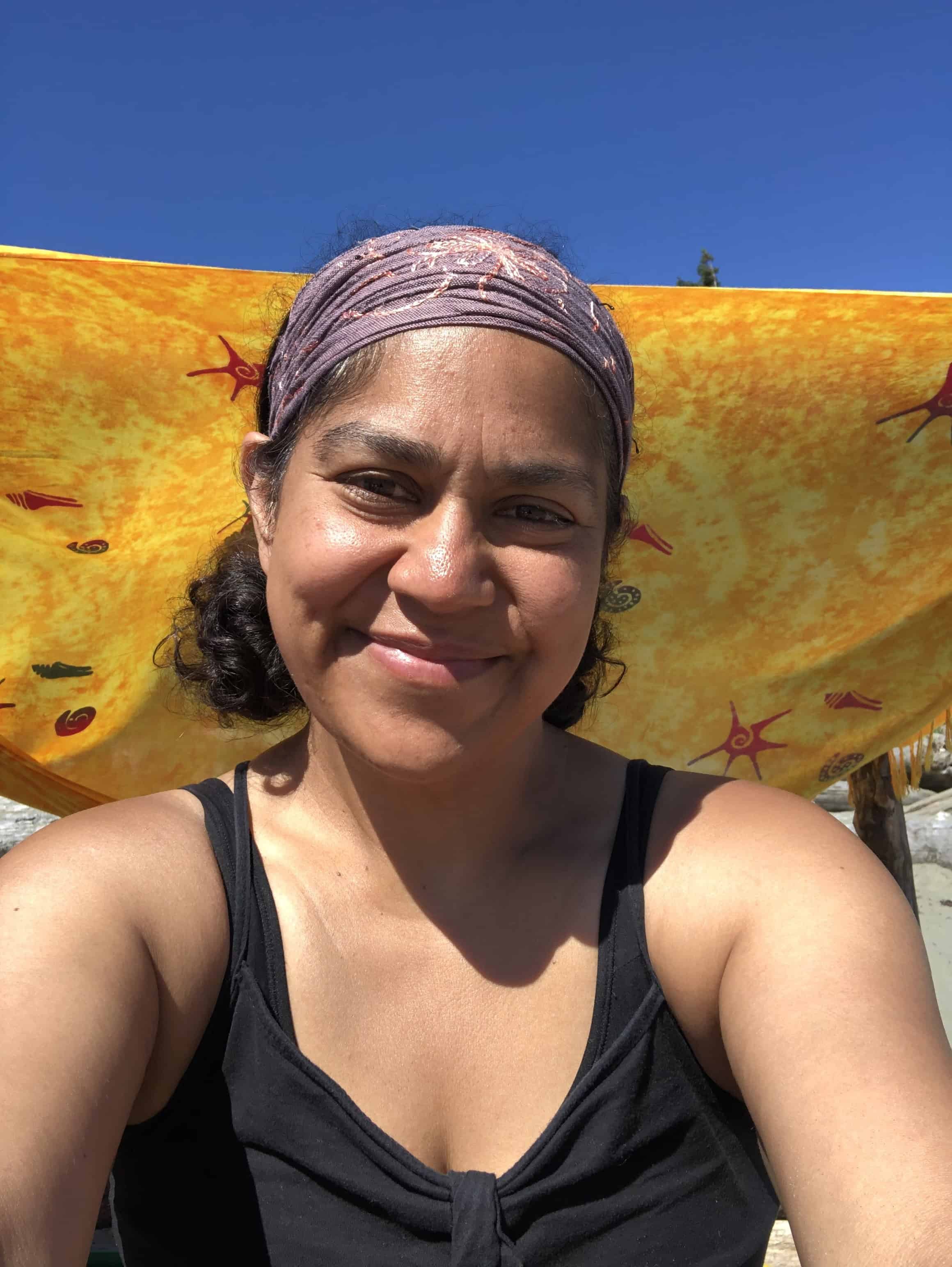Close-up of Natasha Ramlall smiling outdoors in bright sunlight, wearing a patterned headscarf with a vivid yellow-orange fabric backdrop behind her.
