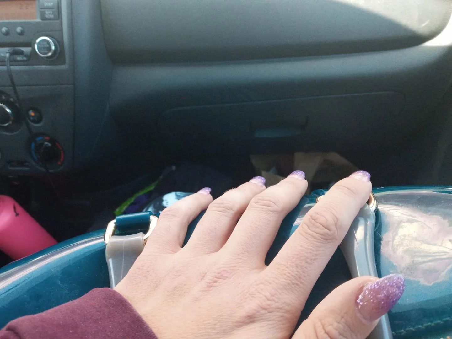 Close-up photo of a hand with glittery purple nail polish resting on a teal handbag inside a car, with the dashboard visible in the background.