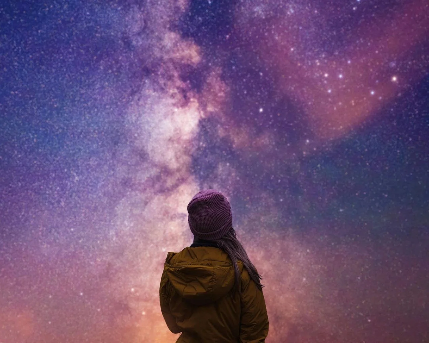 Girl in orange jacket with purple beanie hat looks at the dark night sky with stars, purples, blues, and oranges glowing in the milky way.