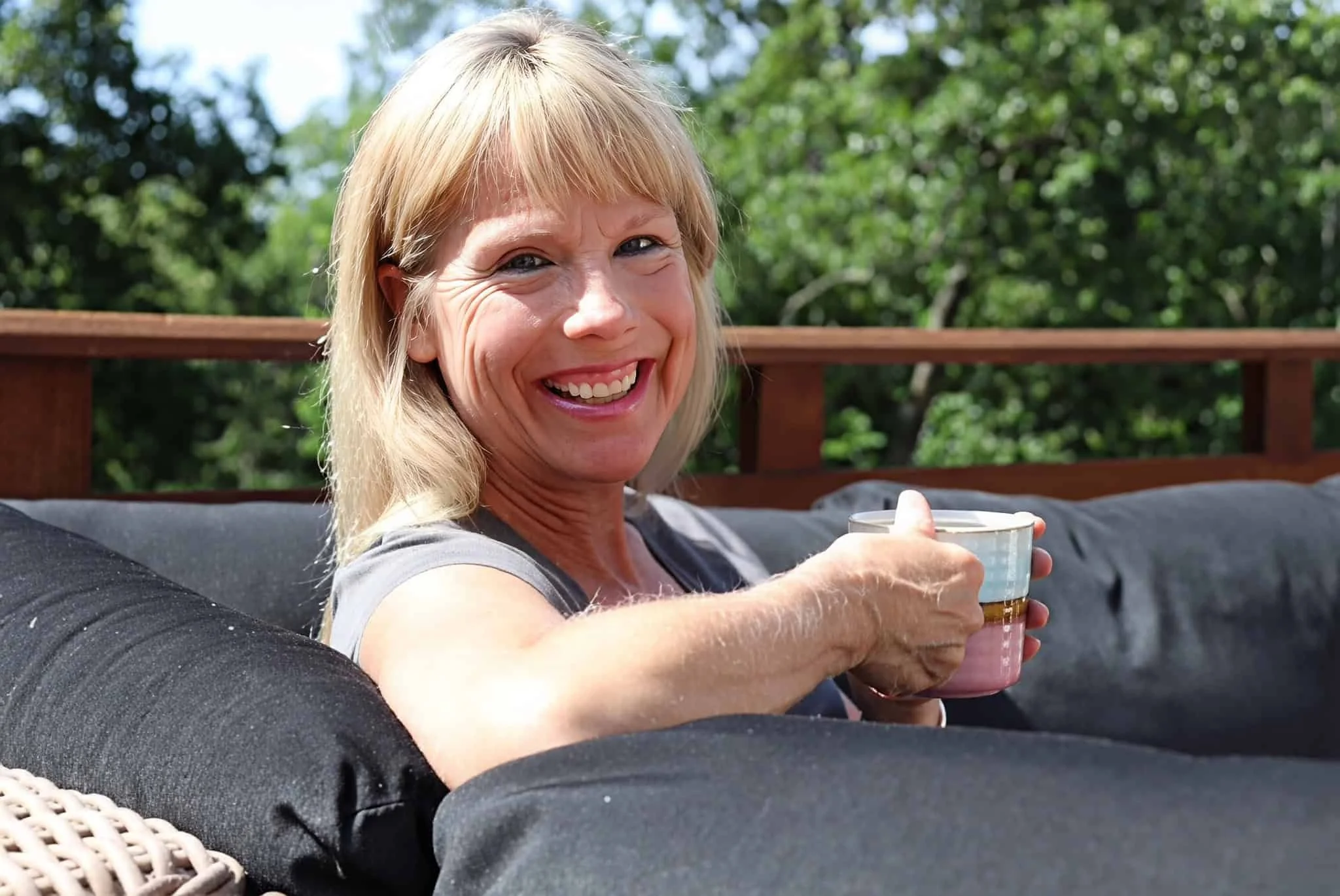 A smiling woman with blonde hair sits on an outdoor couch holding a cup of coffee, with trees and sunlight in the background.