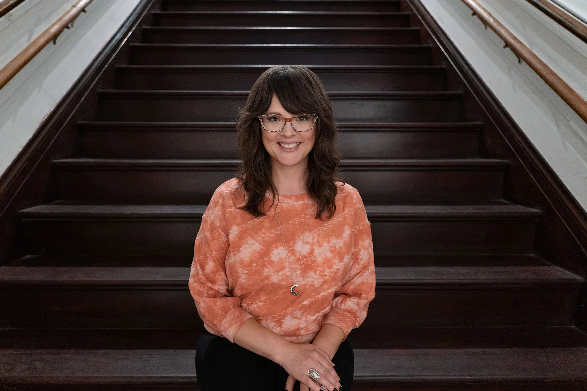 Woman with dark hair and glasses smiling while seated on a wide wooden staircase, wearing an orange tie-dye sweatshirt and a crescent moon necklace.