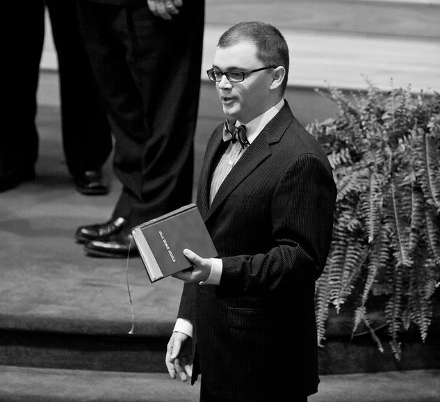 A man in a suit and bow tie speaking from the front of a church sanctuary while holding a Bible during a sermon.