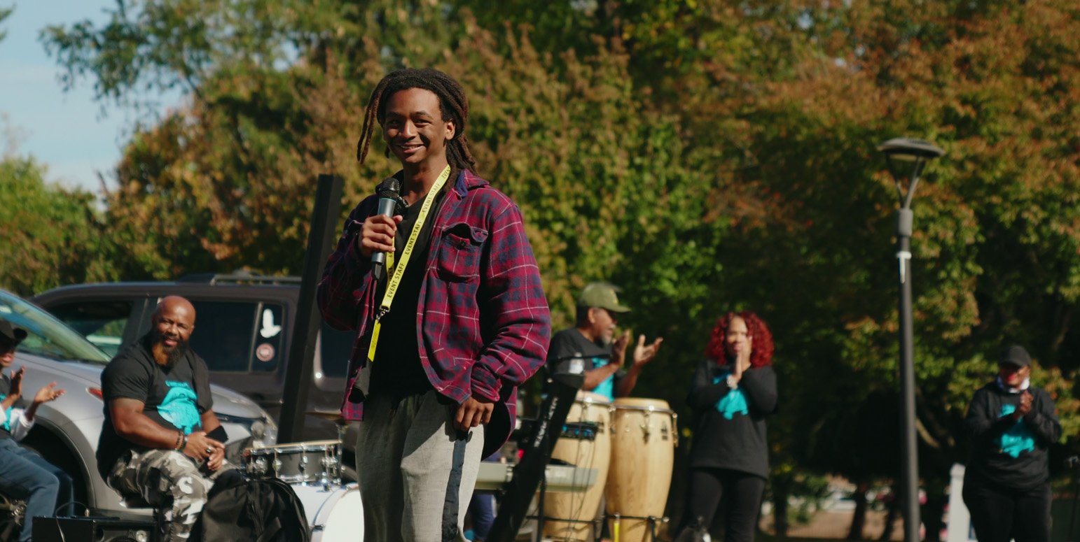 Lawrence C. Harris speaking into a microphone at an outdoor event, with people and instruments in the background
