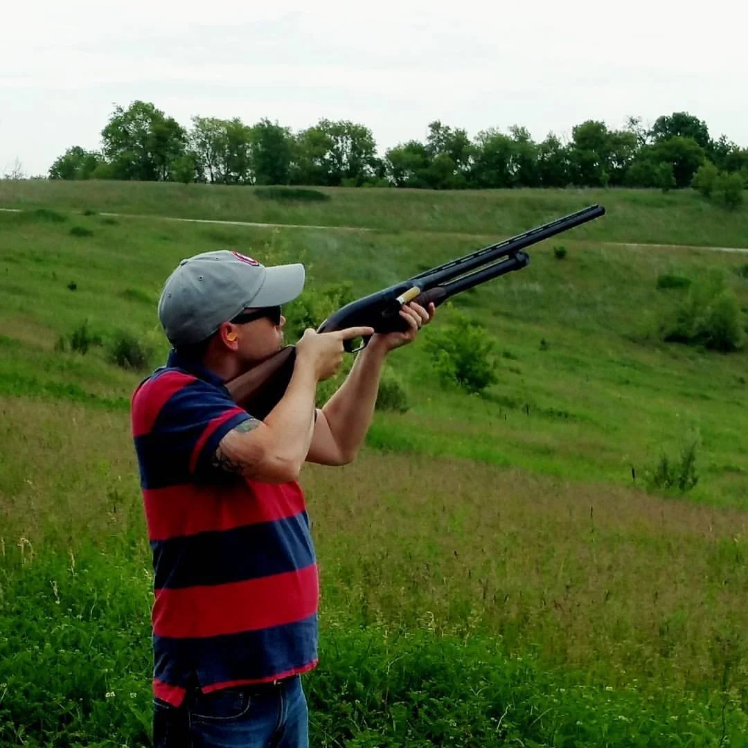 Person participating in skeet shooting at an outdoor range, aiming a shotgun toward the sky in a wide grassy landscape with rolling hills and trees in the background.