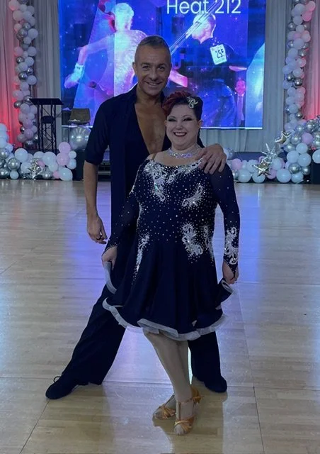 Ballroom dancer in a navy dress with silver detailing poses with her dance partner on a ballroom floor beneath blue event lighting.