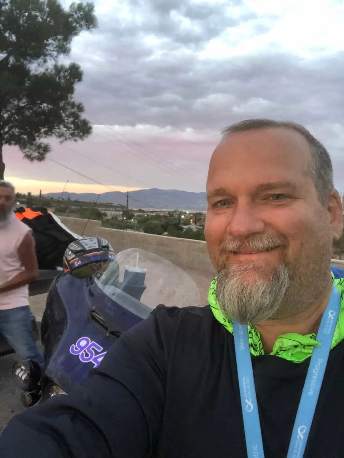 Gray-haired man with a goatee smiling in a selfie with a medal and motorcycle behind him.