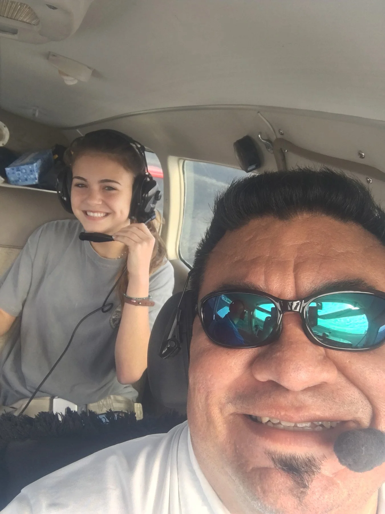 Smiling man and teenage girl wearing aviation headsets inside a small aircraft cockpit during a flight.