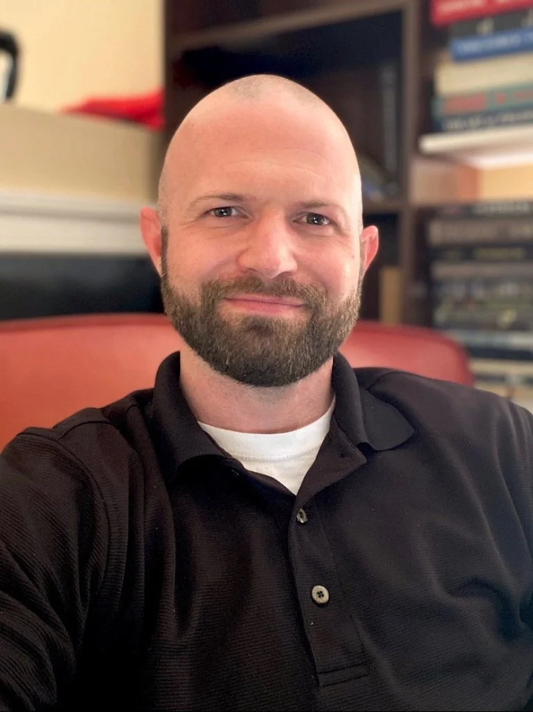 Portrait of a smiling man seated in front of a bookshelf in an office setting.