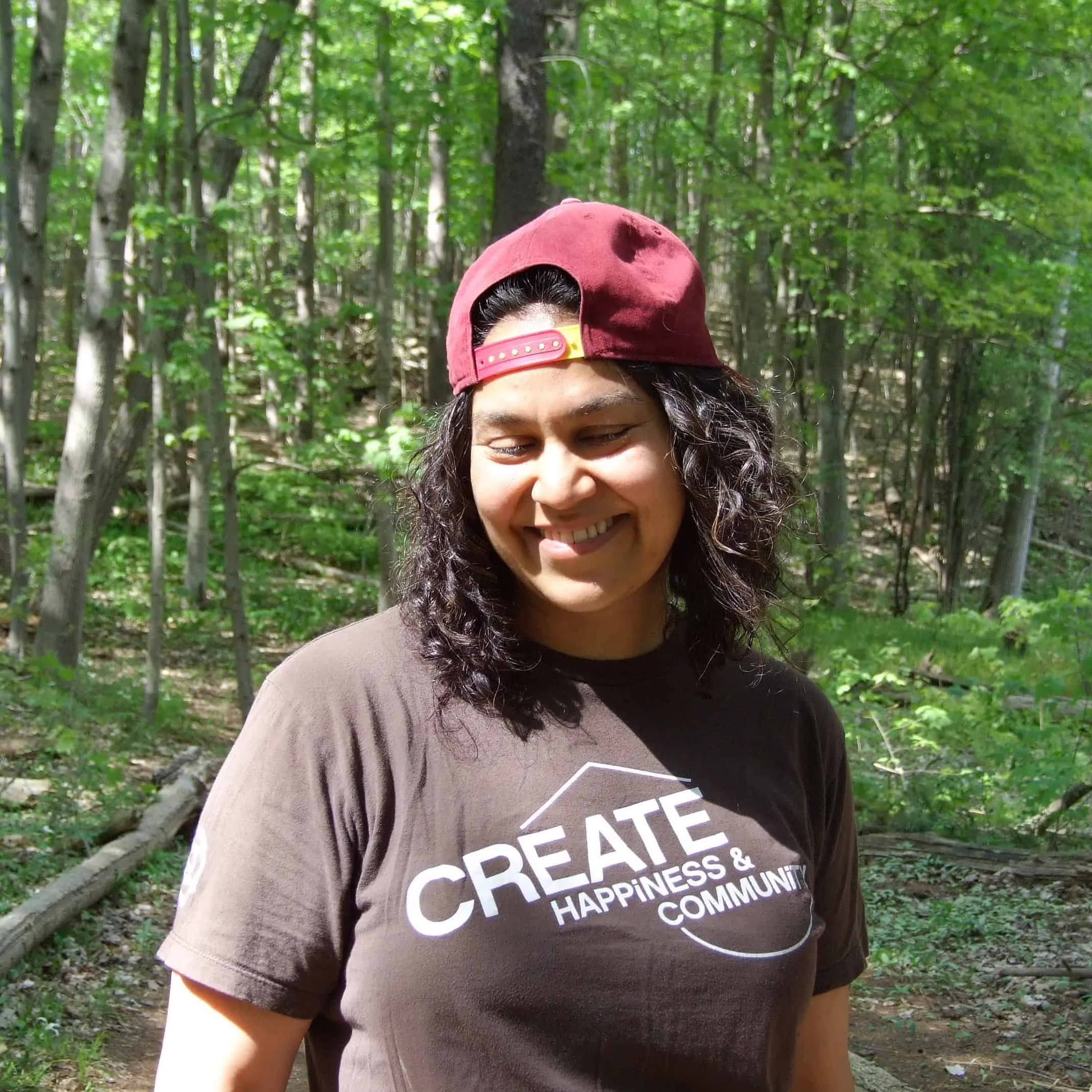 Natasha Ramlall smiling with her eyes closed in a sunlit forest, wearing a backward red cap and a brown T-shirt.