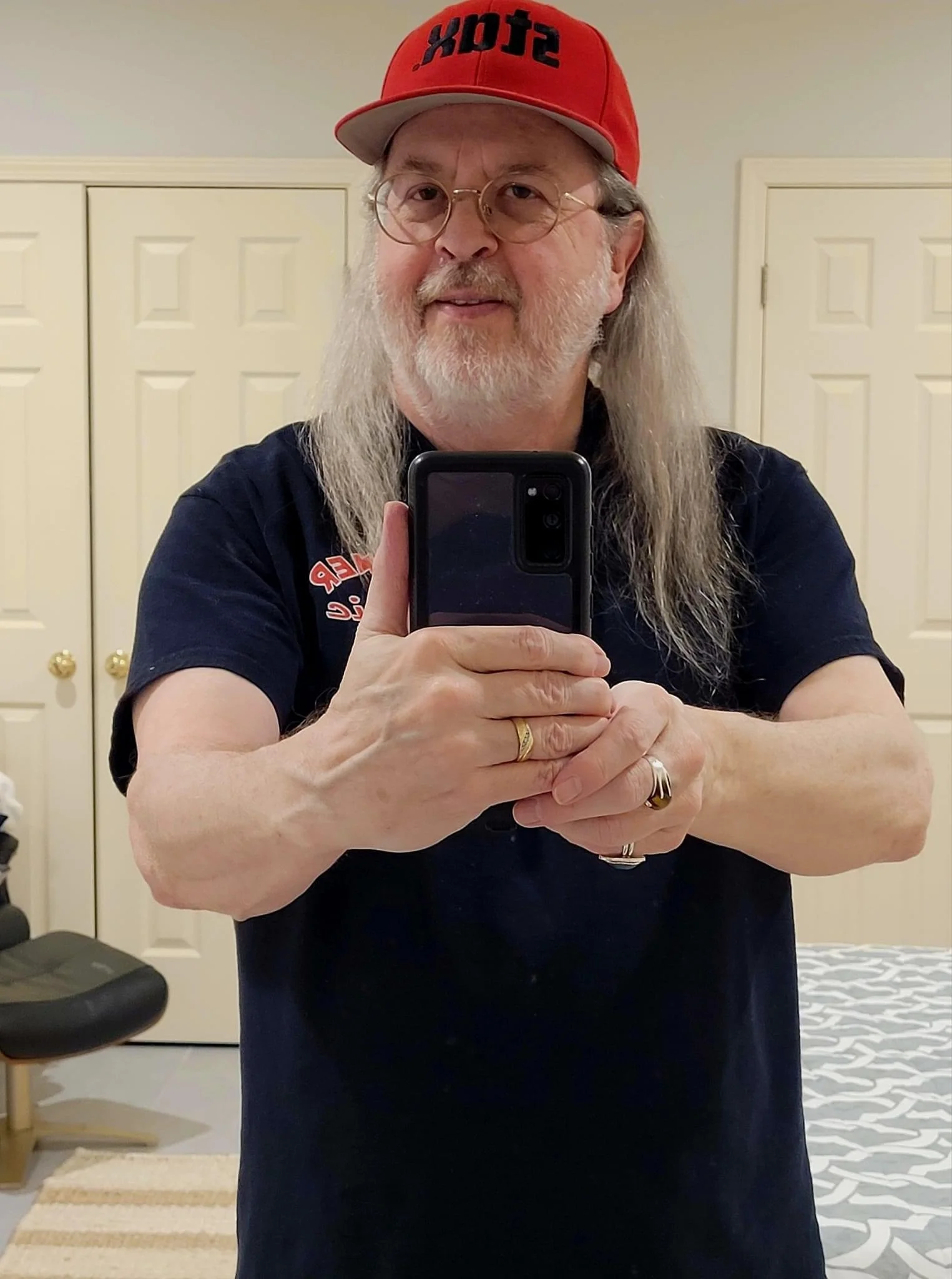 man in his 60s with long hair and a short white beard wears a red hat and poses for a mirror selfie holding his phone in his hand