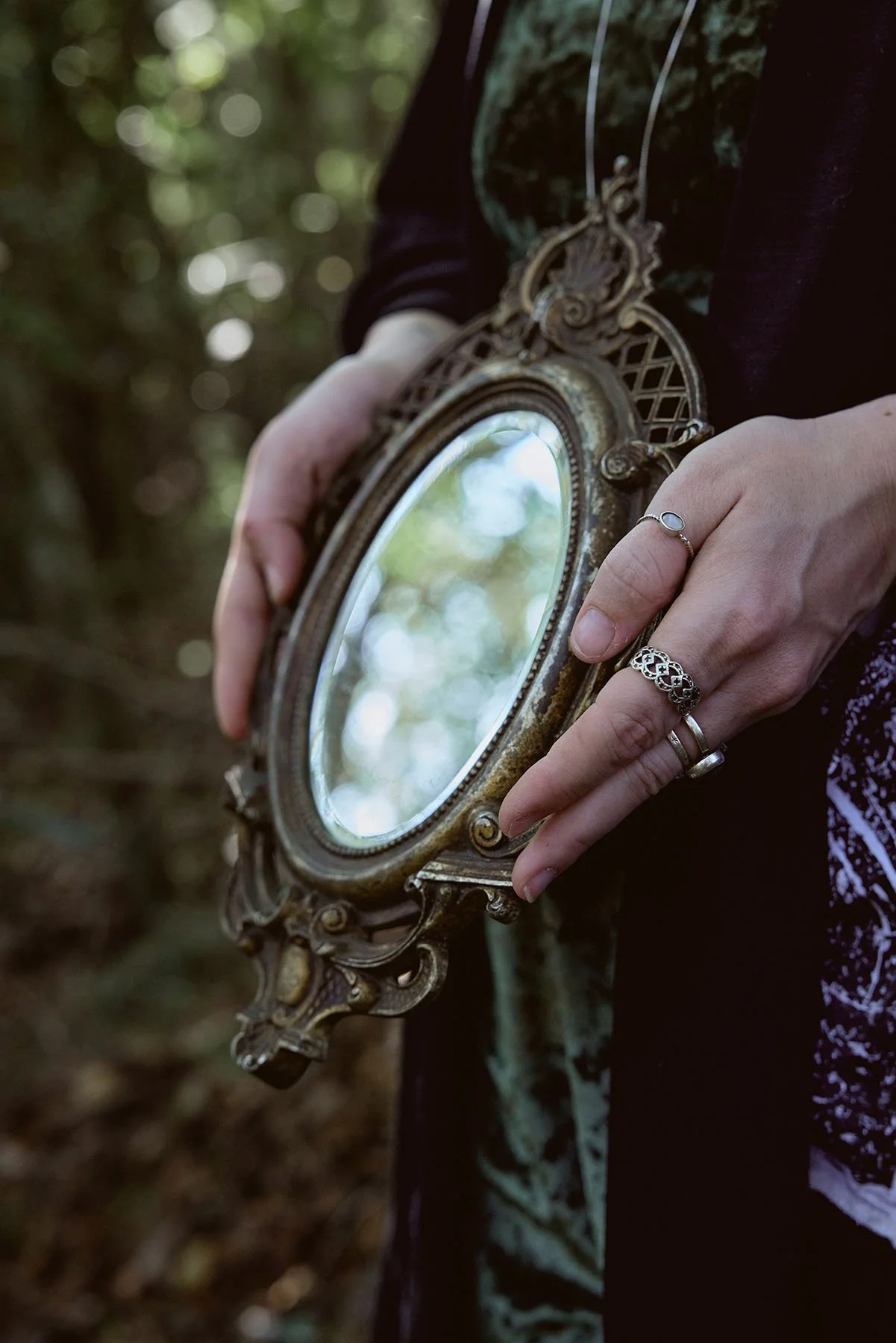 Close-up of hands holding an ornate antique mirror in a wooded area, with sunlight reflecting off the glass and rings visible on the fingers.