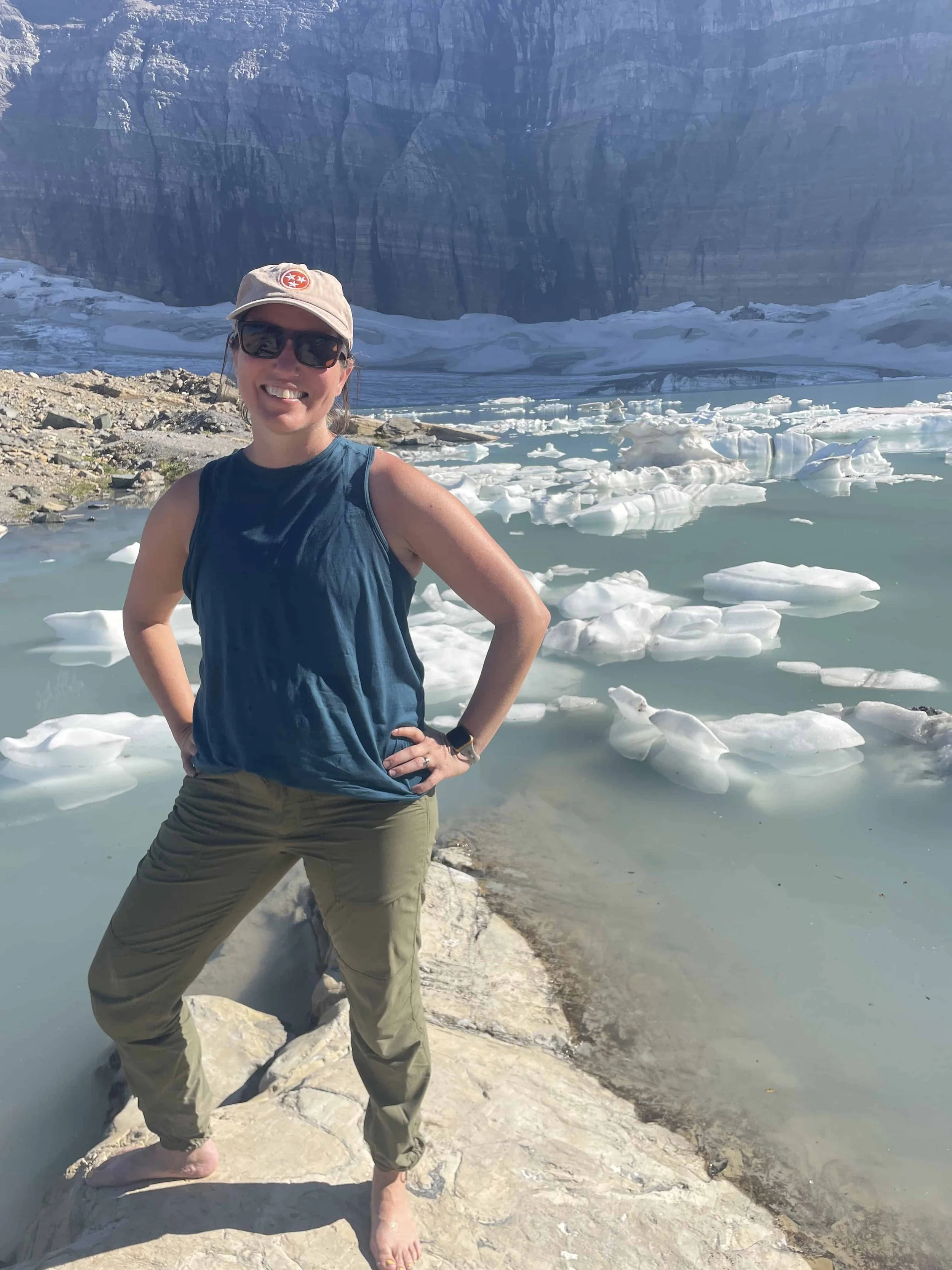 Woman in a baseball cap and sunglasses smiling while posing barefoot on a rock in front of melting ice.