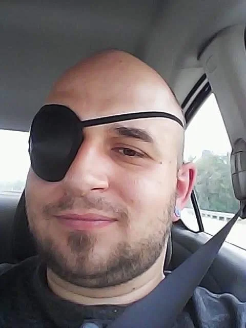 A smiling man wearing an eye patch sits in the front passenger seat of a car with a seatbelt fastened.