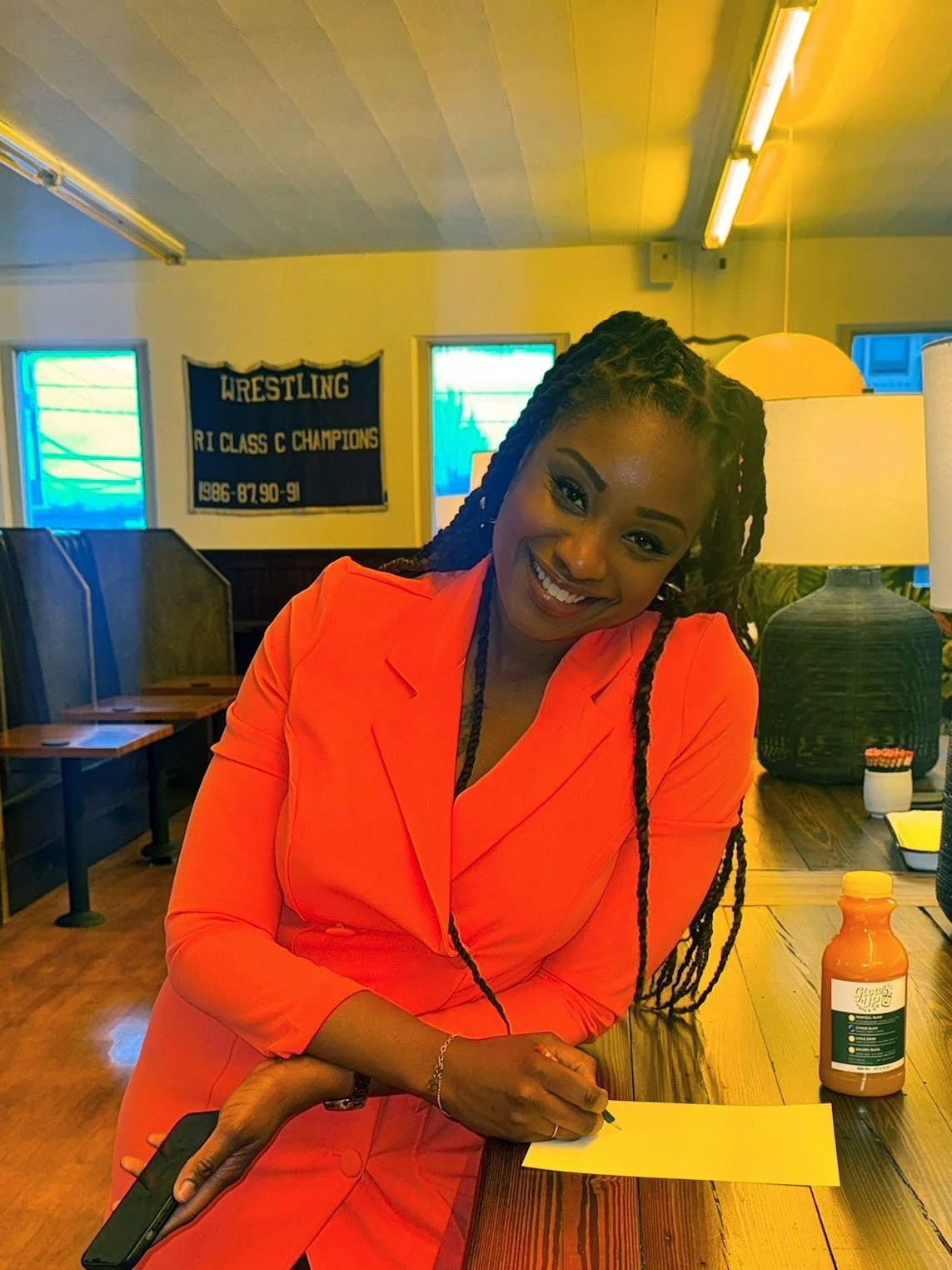 A smiling woman with long braids, wearing a bright coral pantsuit, sits at a wooden table writing on a piece of paper with a bottled Glow Up Juice Co. drink beside her.