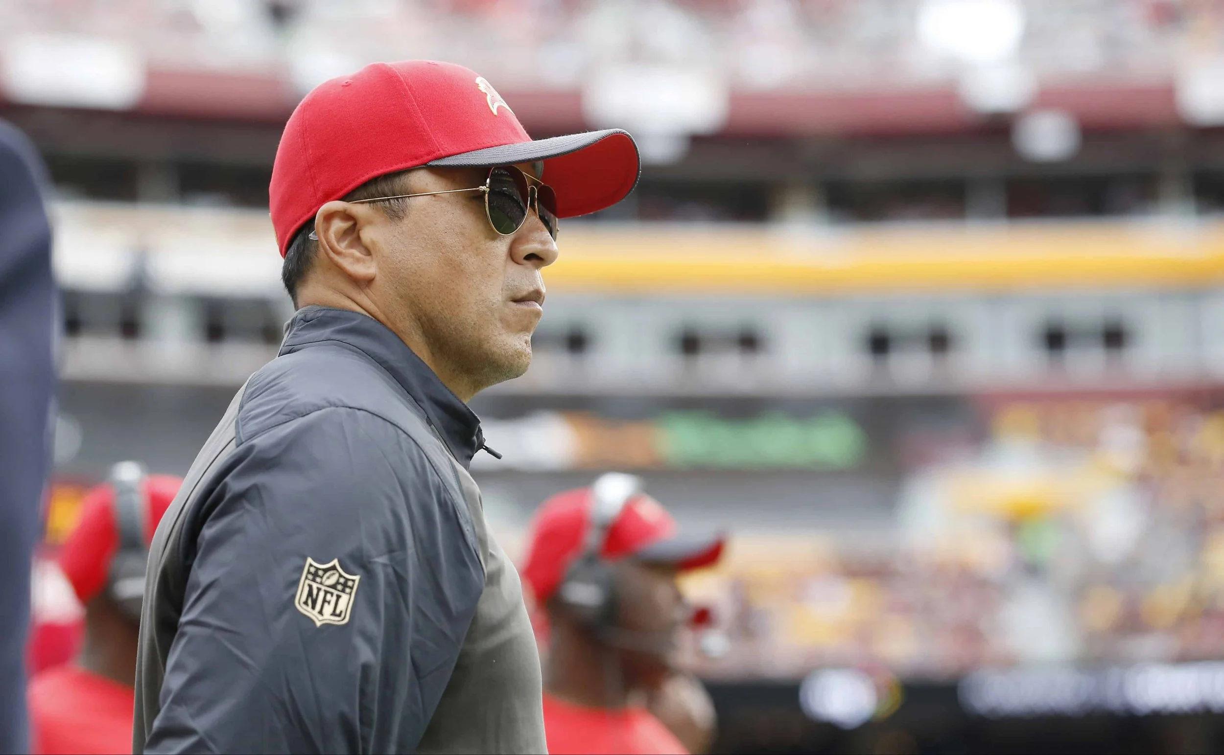 Side profile of a man wearing a red cap, sunglasses, and a dark jacket standing on a football sideline, with a blurred stadium and team staff in the background.