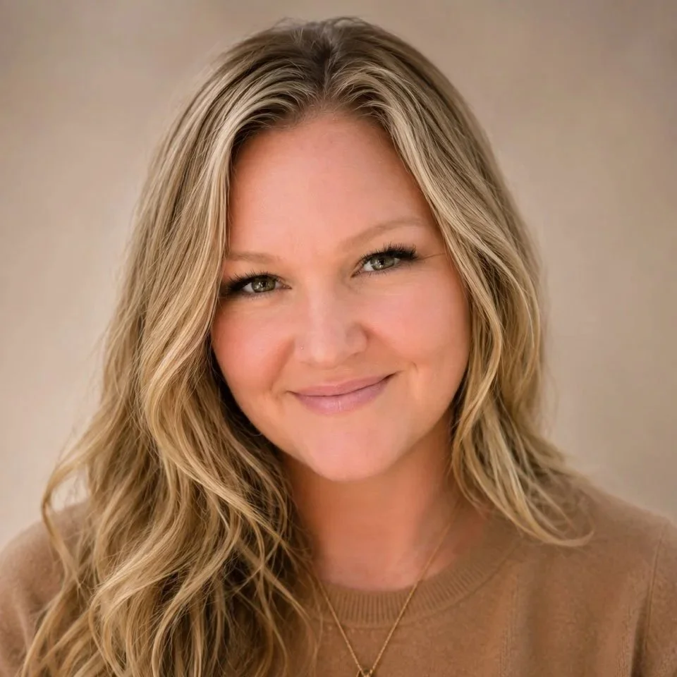 Smiling woman with long blonde hair wearing a tan sweater and pendant necklace against a neutral background.