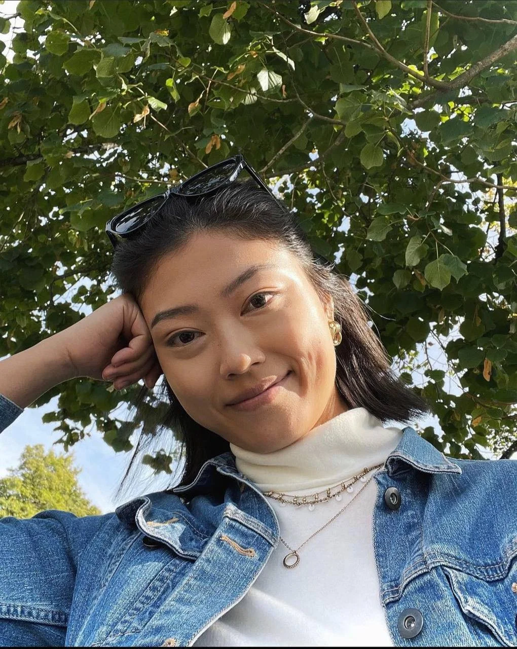 Smiling Asian woman poses in front of a tree looking down at camera with her hand resting on her face