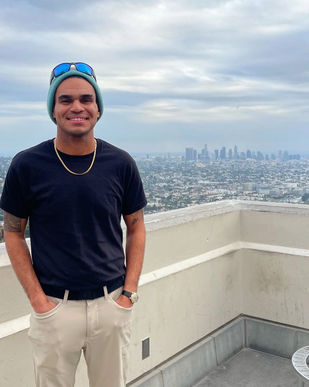 Ty Briggs stands on a rooftop overlooking the Los Angeles skyline on a cloudy day, wearing a black t-shirt, beanie, sunglasses, and gold chain while smiling toward the camera.