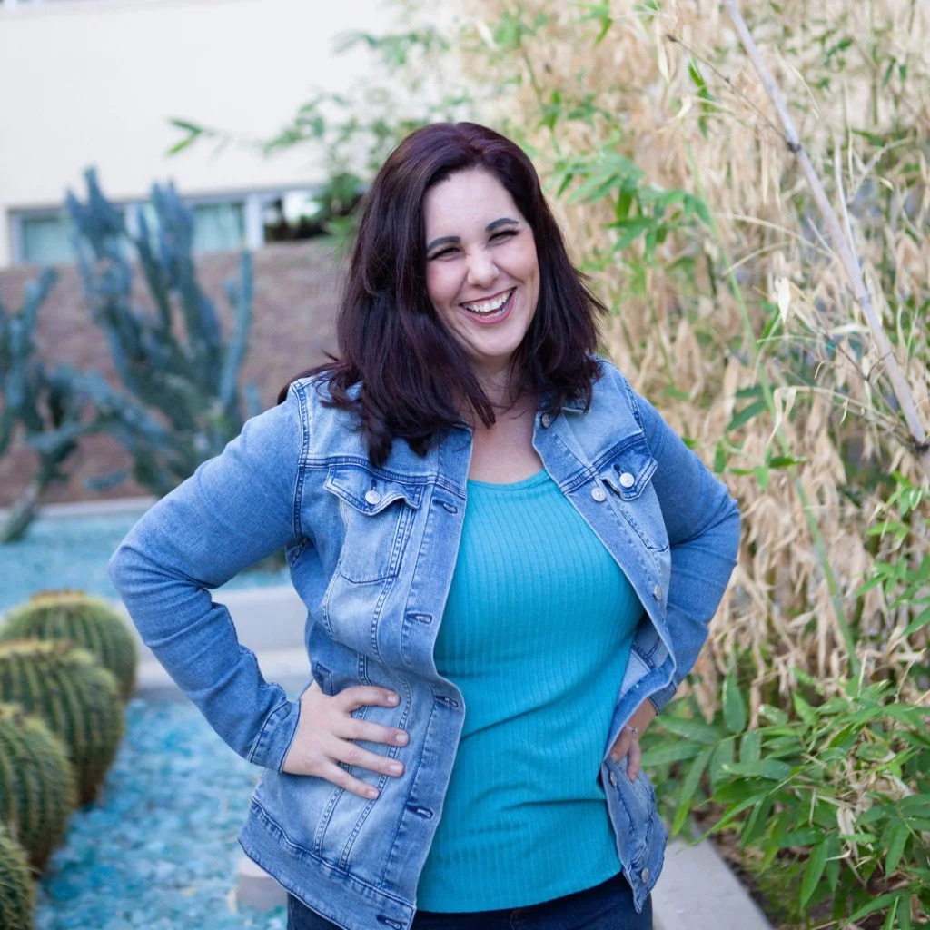 Portrait of a coach, speaker, and mother smiling outdoors in a denim jacket near desert landscaping.