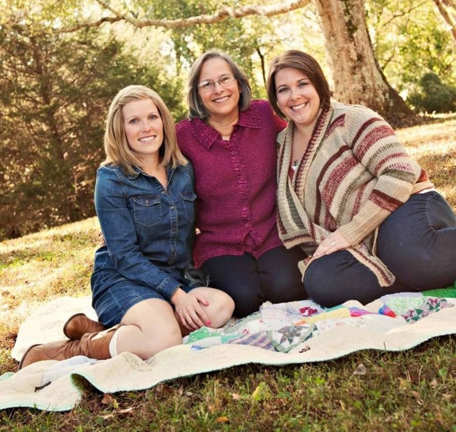 a young woman in a denim dress sits on a blanket outside next to older woman with glasses and burgundy shirt, black pants, with her arms around a woman wearing a striped cardigan