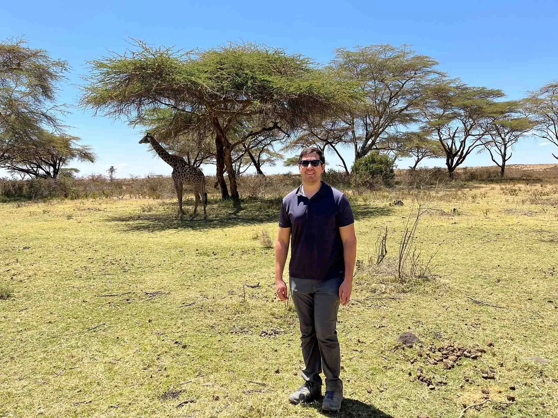 Wesley Warshawer stands in a grassy landscape in Kenya, with acacia trees and a giraffe visible behind him.
