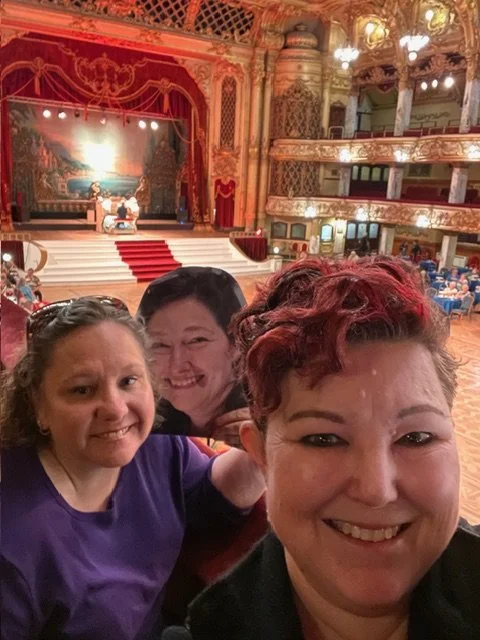 Three smiling women take a selfie inside an ornate theater with red curtains, gold balconies, and audience seating in the background.