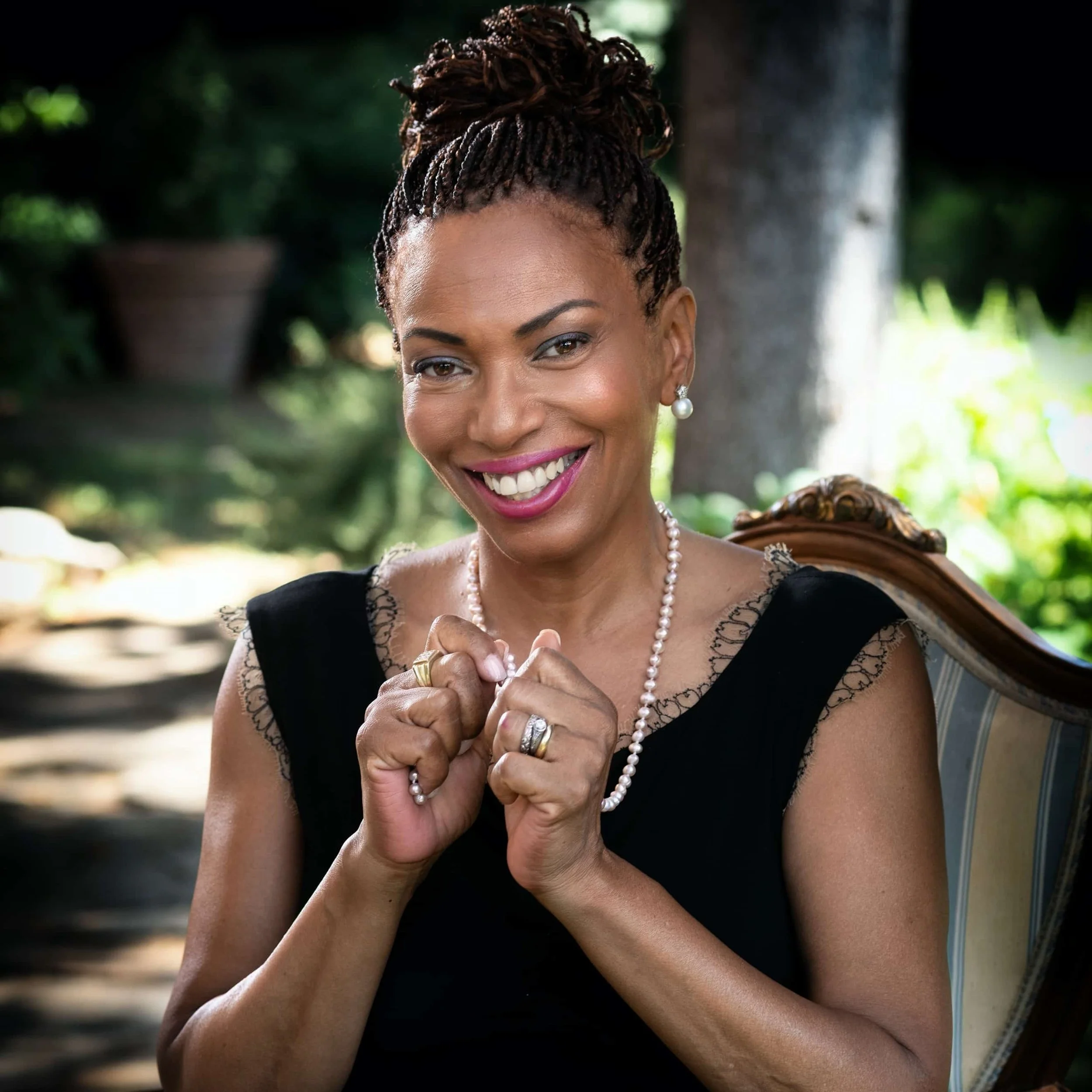 Smiling woman holds her two closed hands together as she poses in a chair.