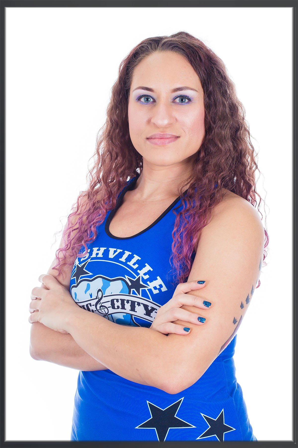 Woman in a blue roller derby jersey stands with her arms crossed against a white background.