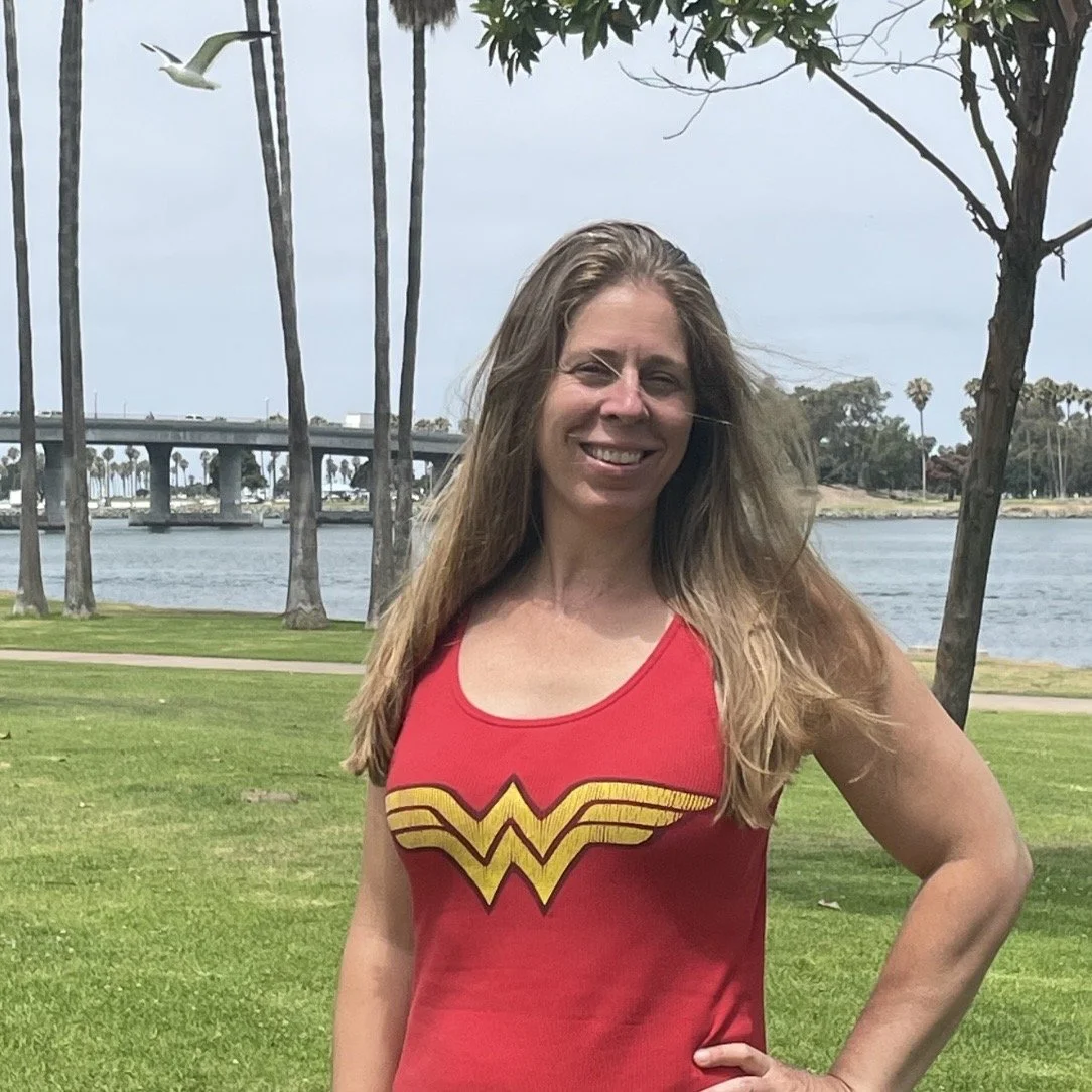 A woman stands outdoors on grass near water, smiling with one hand on her hip, wearing a red Wonder Woman tank top, with palm trees and a bridge in the background.