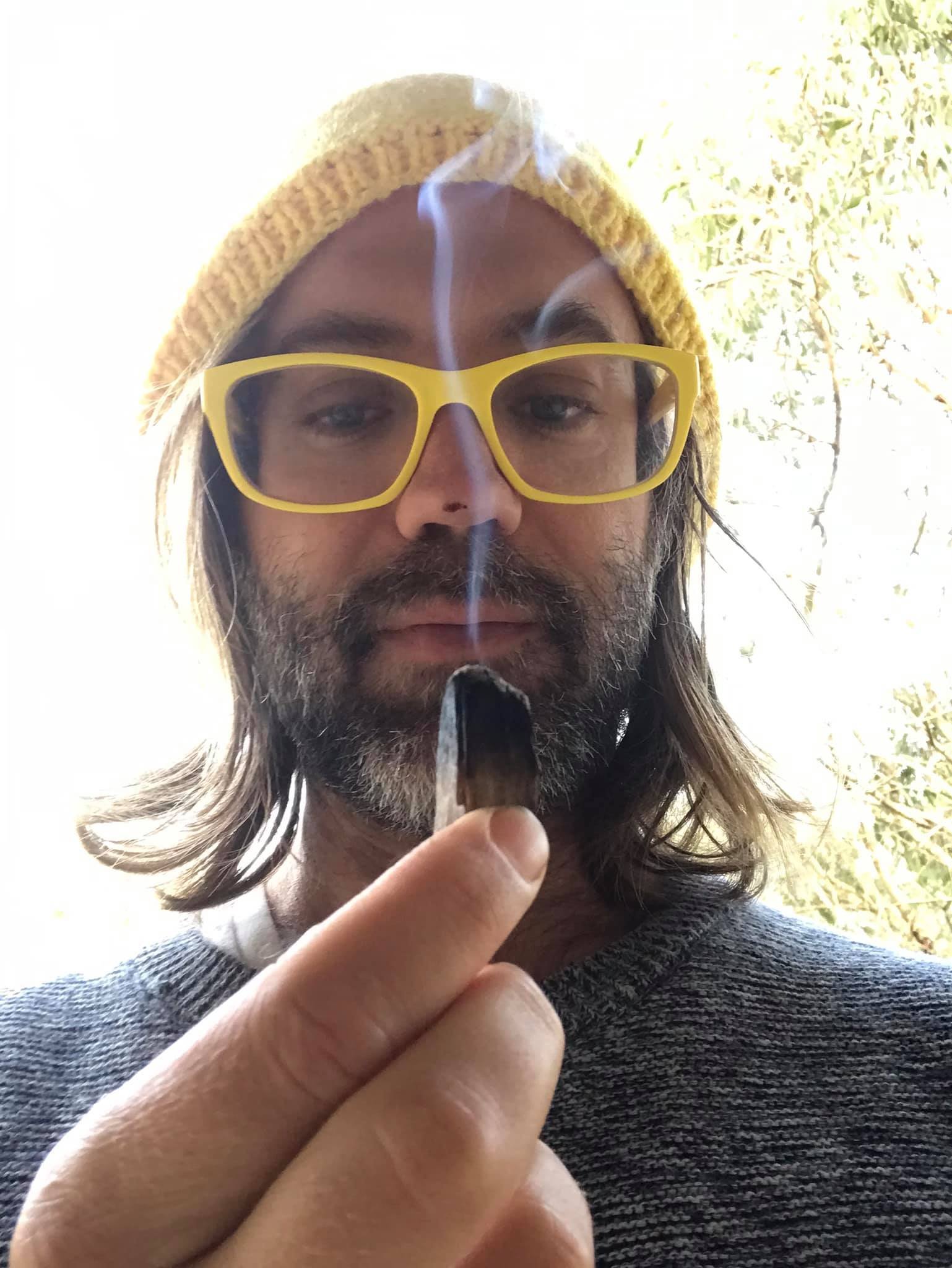 Person outdoors holding a smoking piece of palo santo while wearing yellow glasses and a knit beanie, surrounded by soft natural light and trees.