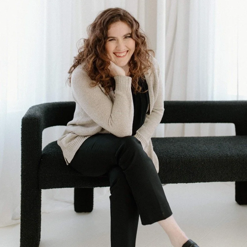 Curly-haired brunette sitting on a bench with head on hand and legs crossed.