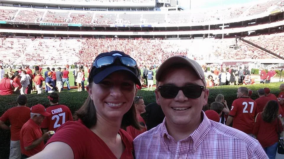 Smiling couple posing for a selfie at a college football game, standing near the field with a packed stadium crowd in the background on a sunny day.