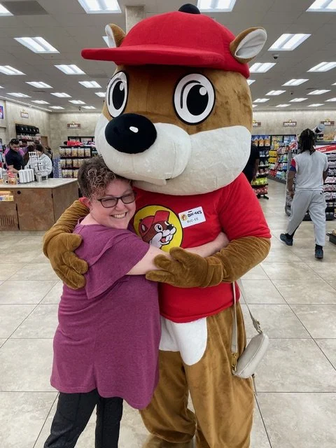 Smiling woman in a mauve shirt hugs the Buc-ee’s beaver mascot inside a brightly lit store.