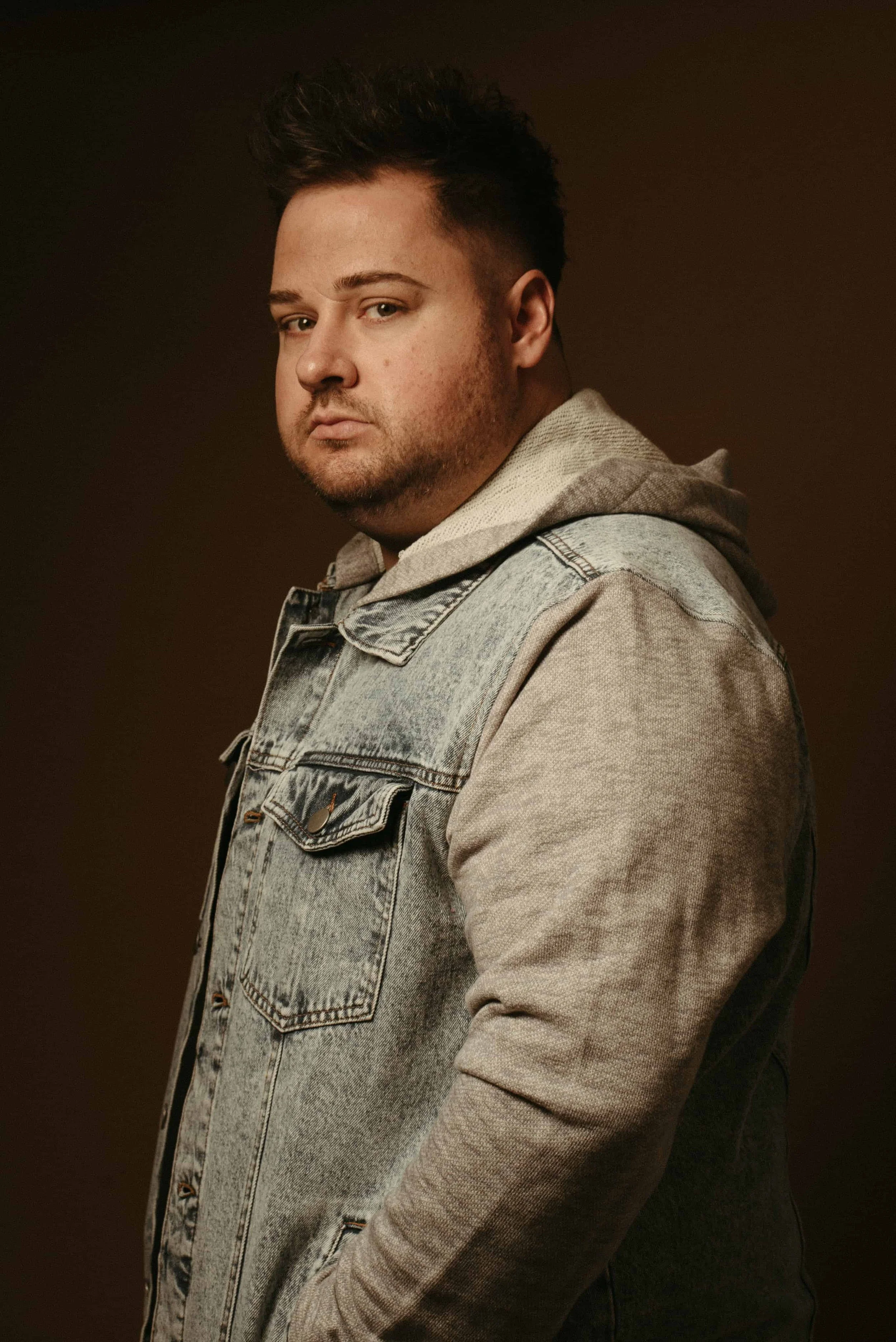 Portrait of a man in a denim jacket layered over a gray hoodie against a dark studio background.