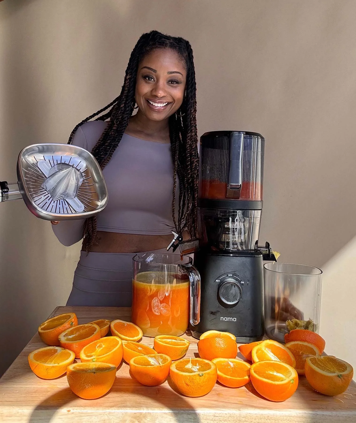 A smiling woman with long braids stands behind a countertop filled with halved oranges, holding part of a juicer beside a pitcher of fresh orange juice and a black Nama juicing machine.