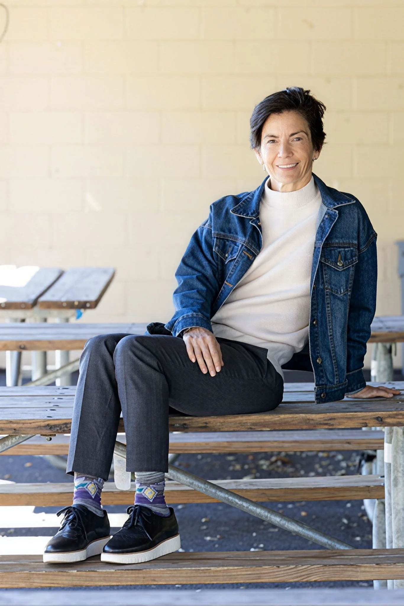 Woman in a denim jacket and gray pants sitting on a wooden picnic table outdoors, smiling at the camera.
