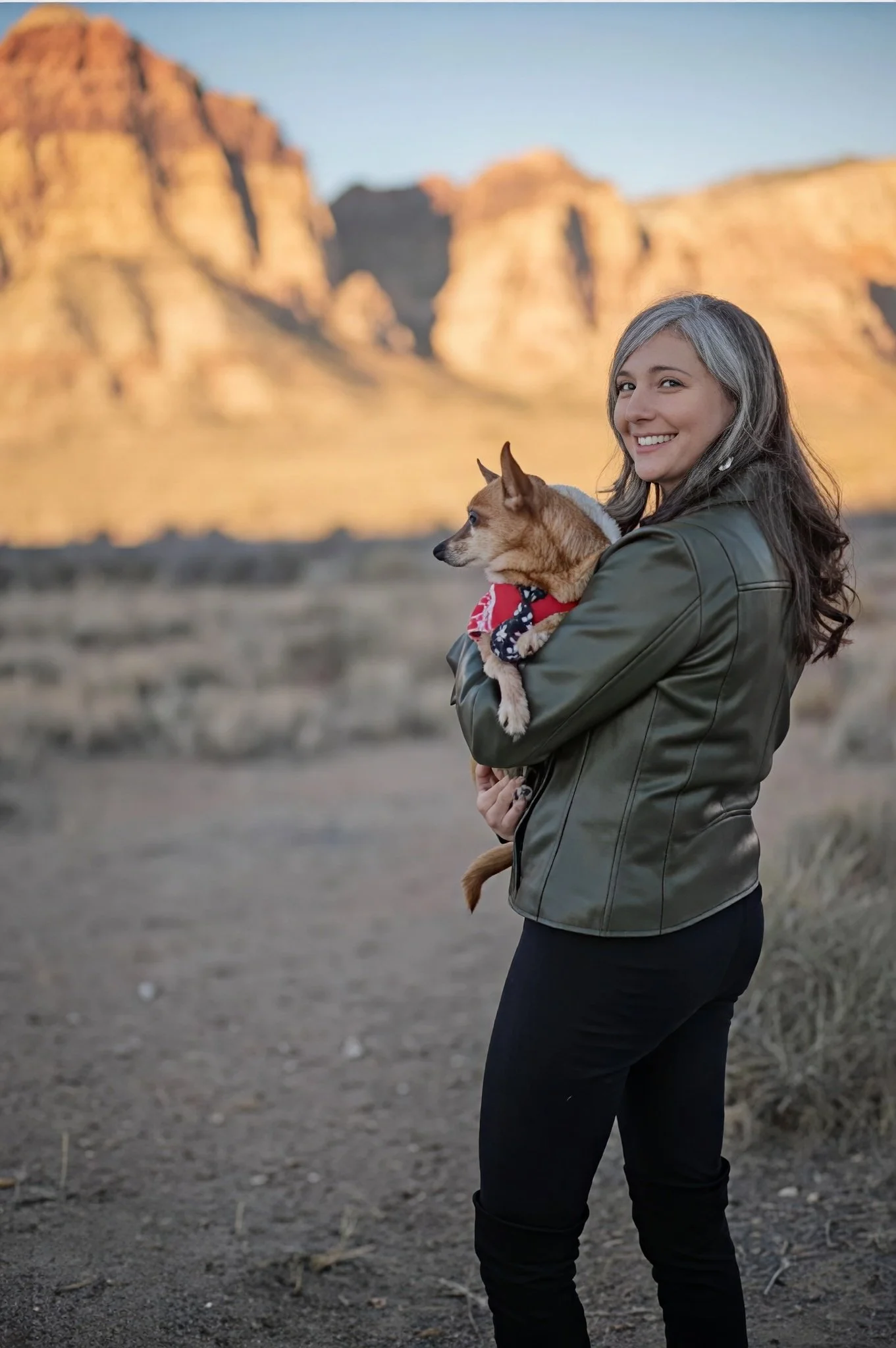 Sheryl Green smiling while holding a small dog in a desert landscape with sunlit mountains blurred in the background. She is wearing a black leather jacket and standing on a dirt path outdoors at sunset.