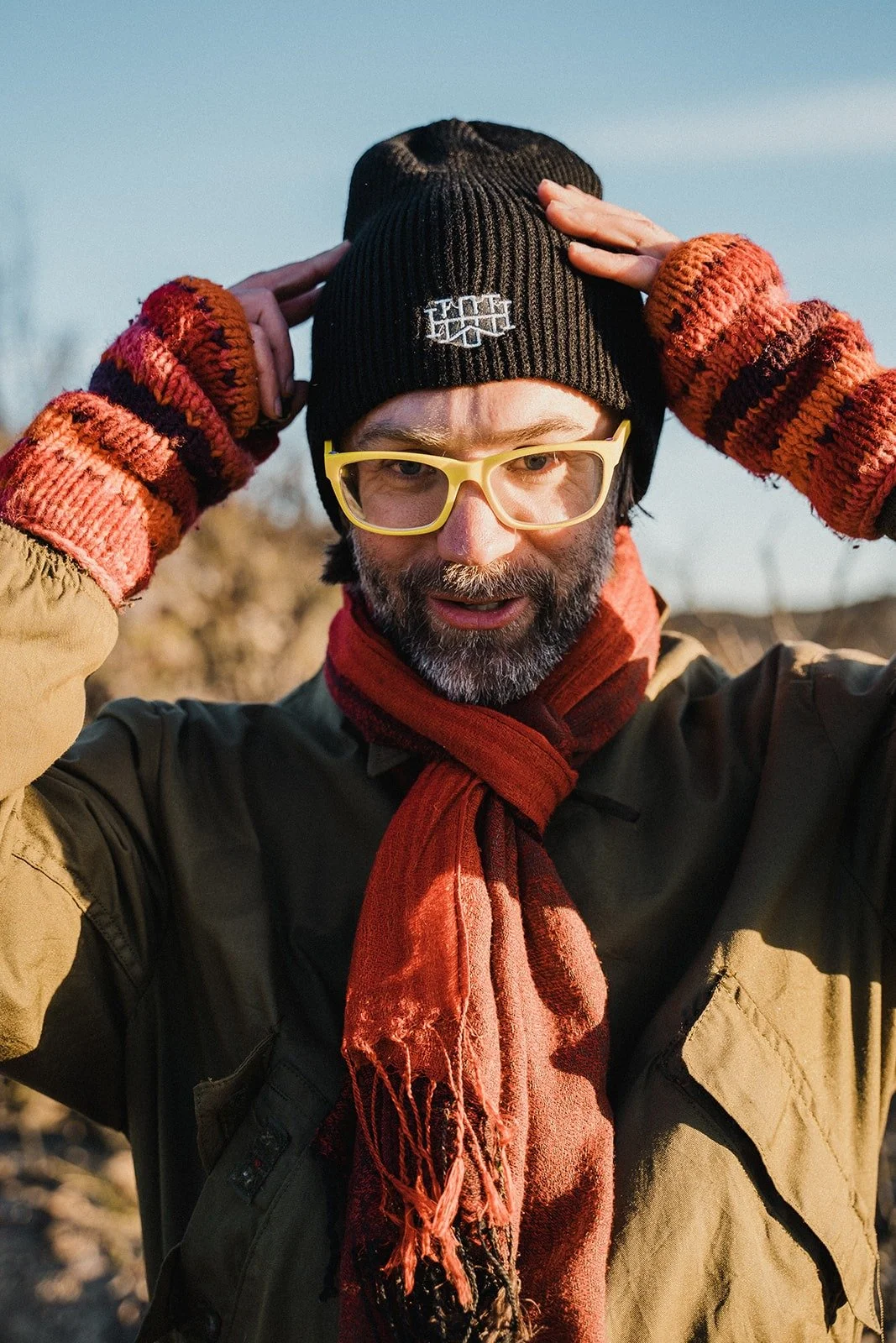 Person wearing a black beanie, yellow glasses, scarf, and colorful knit gloves outdoors in cool weather with sunlight on their face.