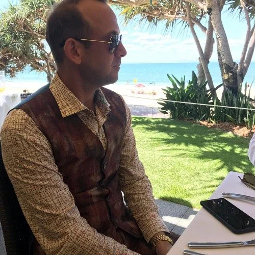 Man wearing sunglasses and a brown vest seated at an outdoor restaurant overlooking the beach and ocean.