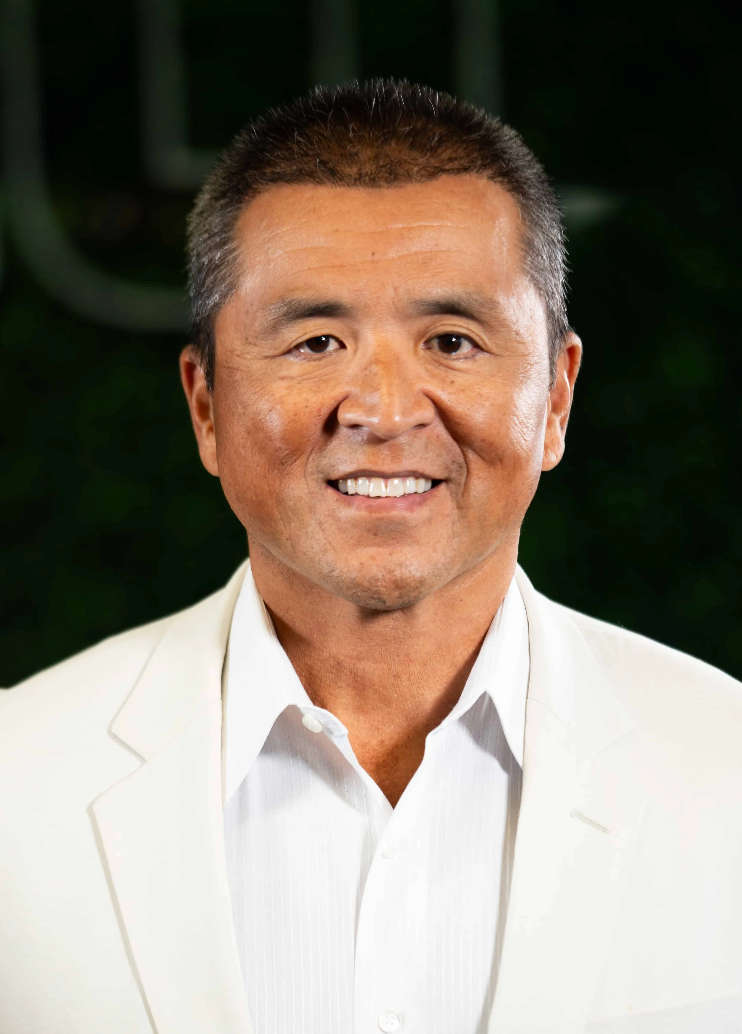 Portrait of a smiling middle-aged man with short dark hair wearing a white jacket and white shirt, facing the camera against a blurred green background.