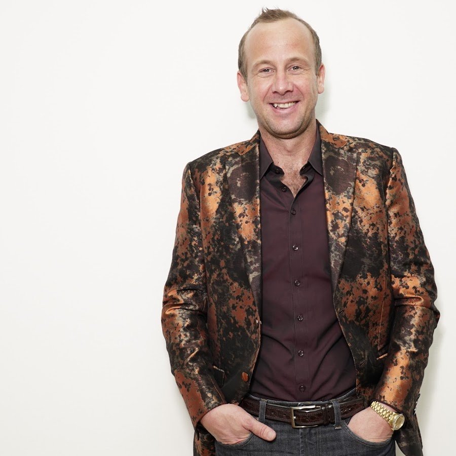 Man smiling in a studio while wearing a shiny orange-and-black patterned suit jacket with his hands in his pockets.