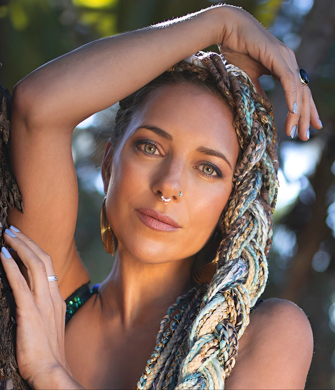Close-up portrait of woman with braided pastel hair, facial piercings, and soft natural makeup posing outdoors beside a tree.
