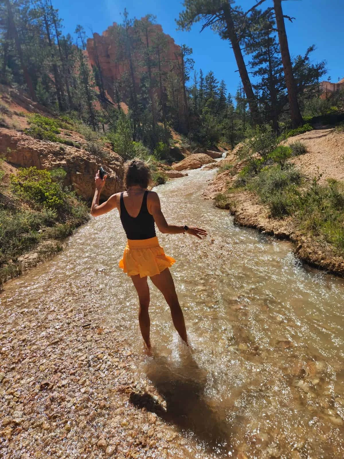 Katie Jensen wades barefoot through a shallow creek in a red rock canyon, surrounded by trees and sunlight, capturing a joyful outdoor adventure moment.