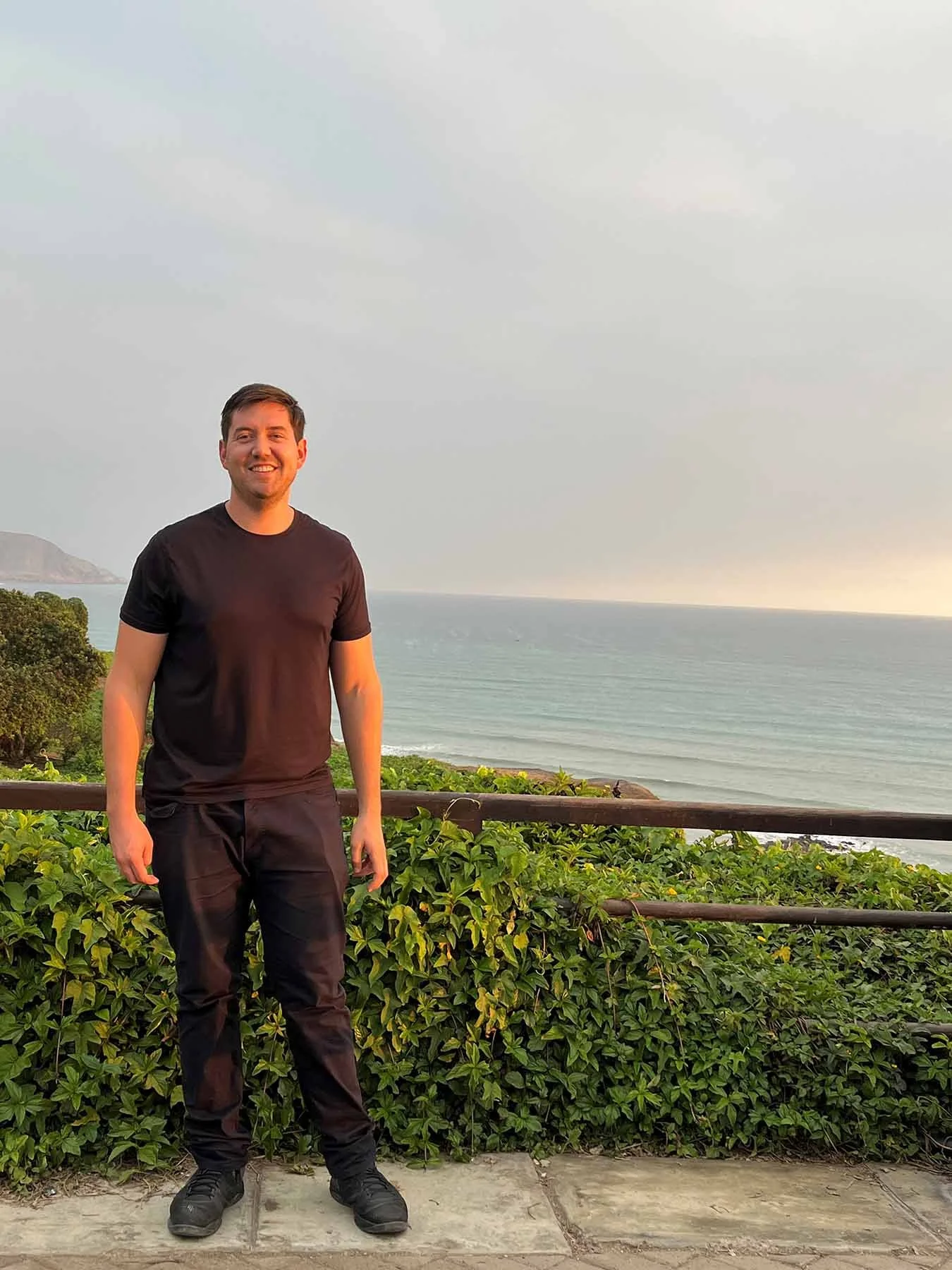 Wesley Warshawer stands smiling at a coastal overlook in Lima, Peru, with the ocean and hazy sky behind him.