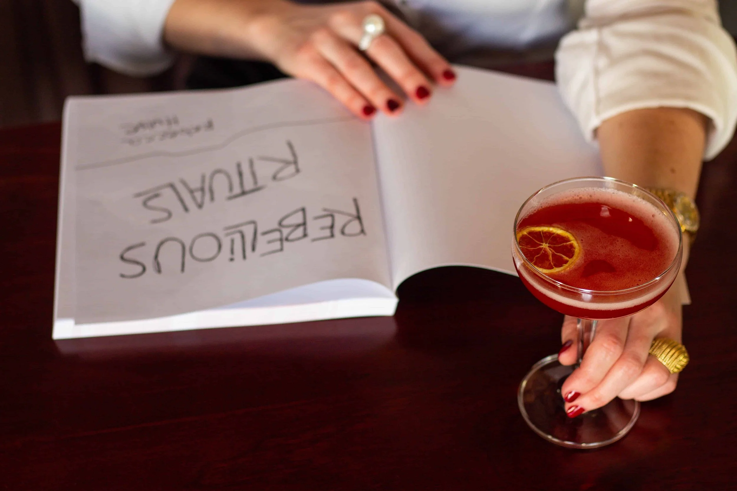 Hands holding an open copy of Rebellious Rituals beside a cocktail drink.