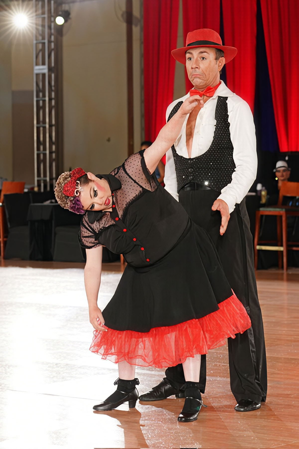 Ballroom dance partners pose in matching black and red costumes, with the woman leaning sideways while her partner stands behind her on the dance floor.