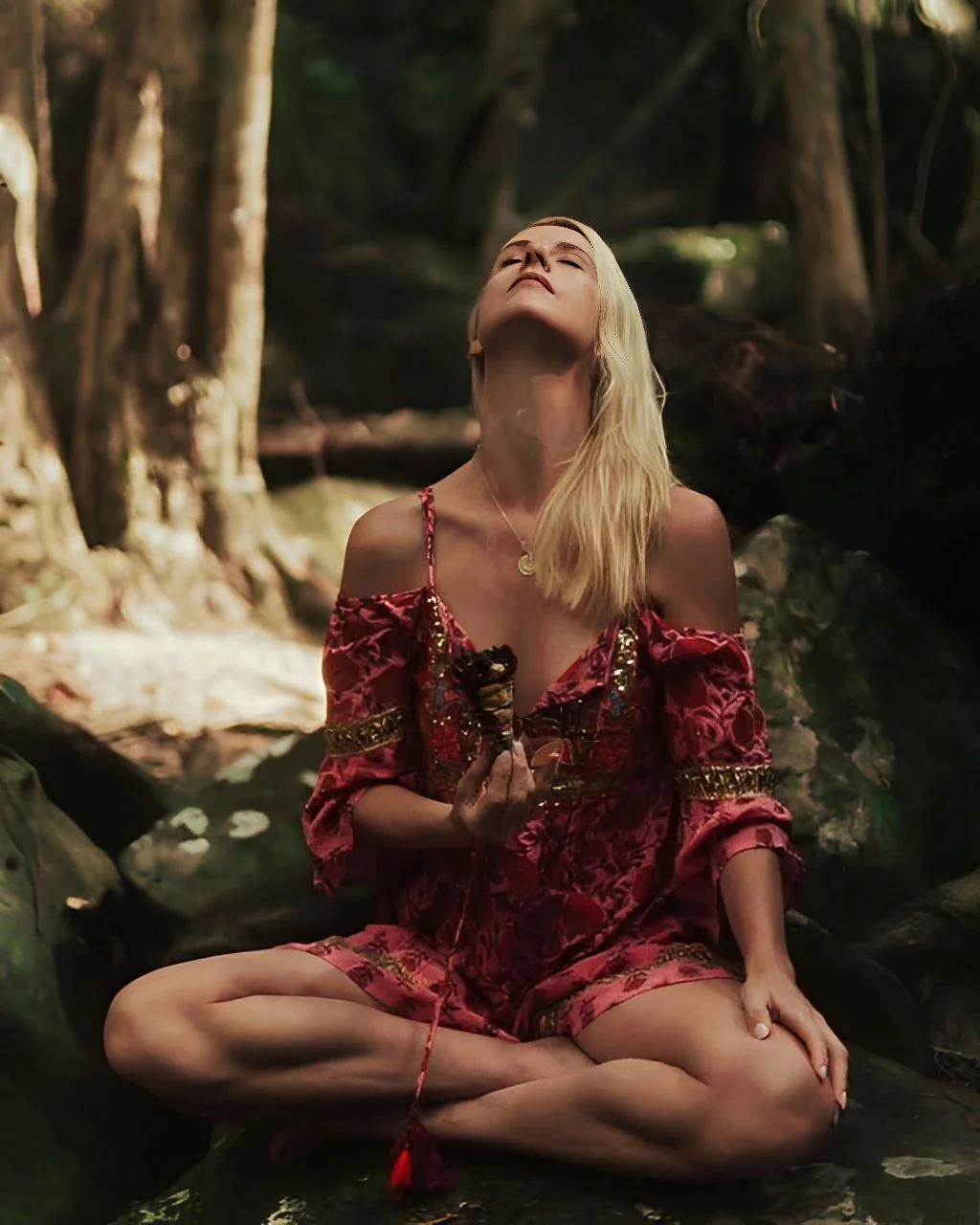 Blonde woman sitting cross-legged on rocks in a forest stream with eyes closed, holding a bundle of herbs or incense during an outdoor spiritual ritual.