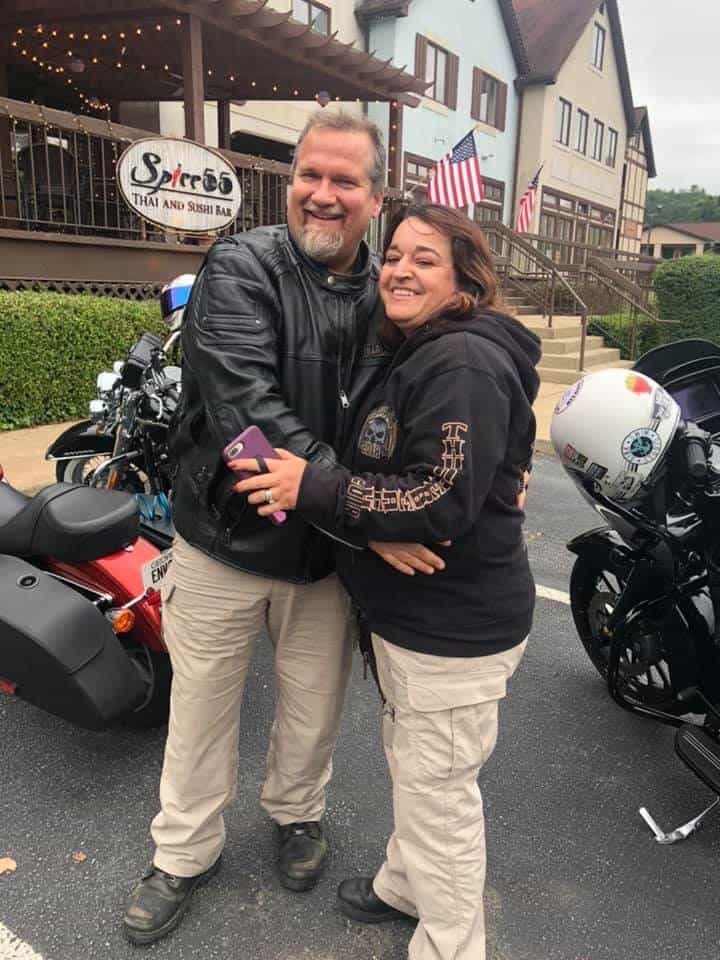 Husband and wife embrace each other, posing for a photo in front of their parked motorcycles.